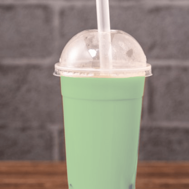 A clear plastic cup filled with green bubble tea, topped with whipped cream and tapioca pearls, sitting on a grey brick surface.